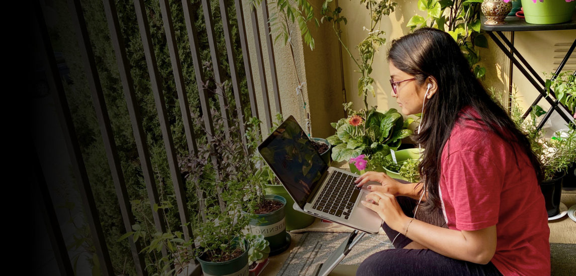 Student sits on her balcony with flowers typing on laptop