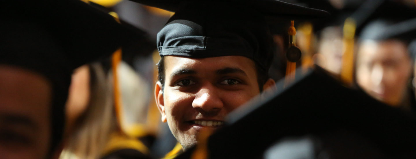 Graduate student in gown and mortarboard hat looks towards camera and smiles among other graduates at the 2022 May CPS Graduation Ceremony. Photo by Heratch Ekmekijan